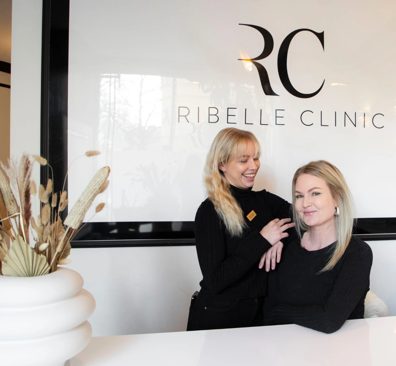 Två medarbetare i svart uniform bakom Ribelle Clinics vita reception med RC-logotypen på väggen