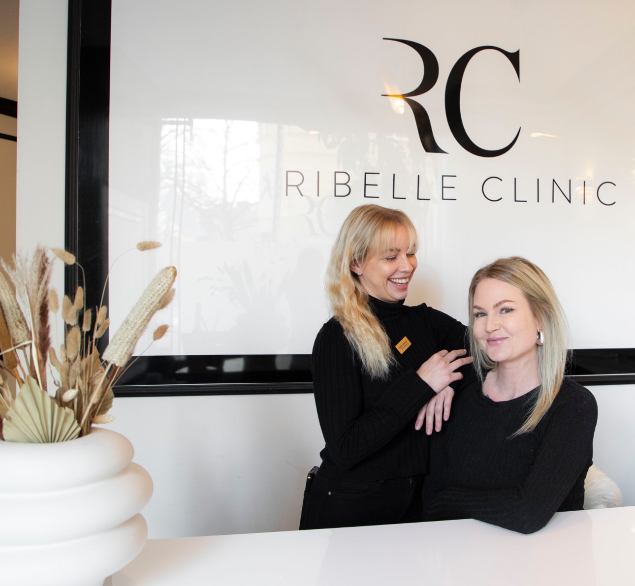 Två medarbetare i svart uniform bakom Ribelle Clinics vita reception med RC-logotypen på väggen